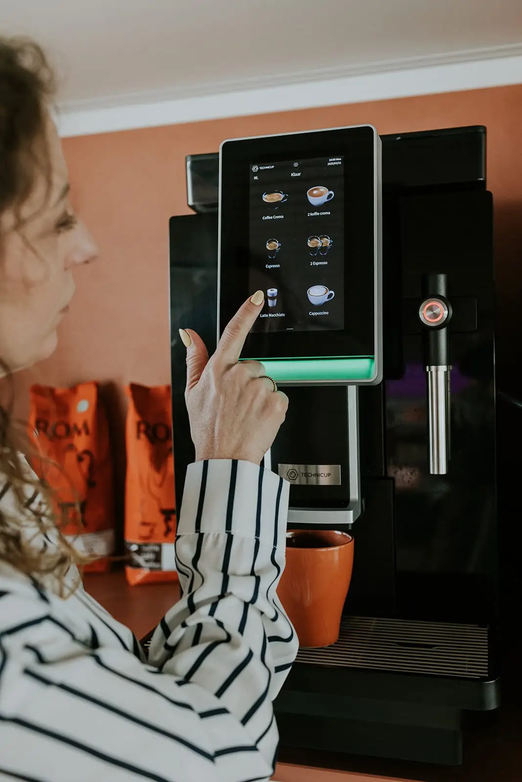 Medewerkster bedient het touchscreen van een professionele koffiemachine, met ROM-koffiezakken ernaast